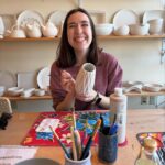 A person enjoying a pottery painting workshop, featuring various ceramic vases, bowls, and paints on the shelves and table.